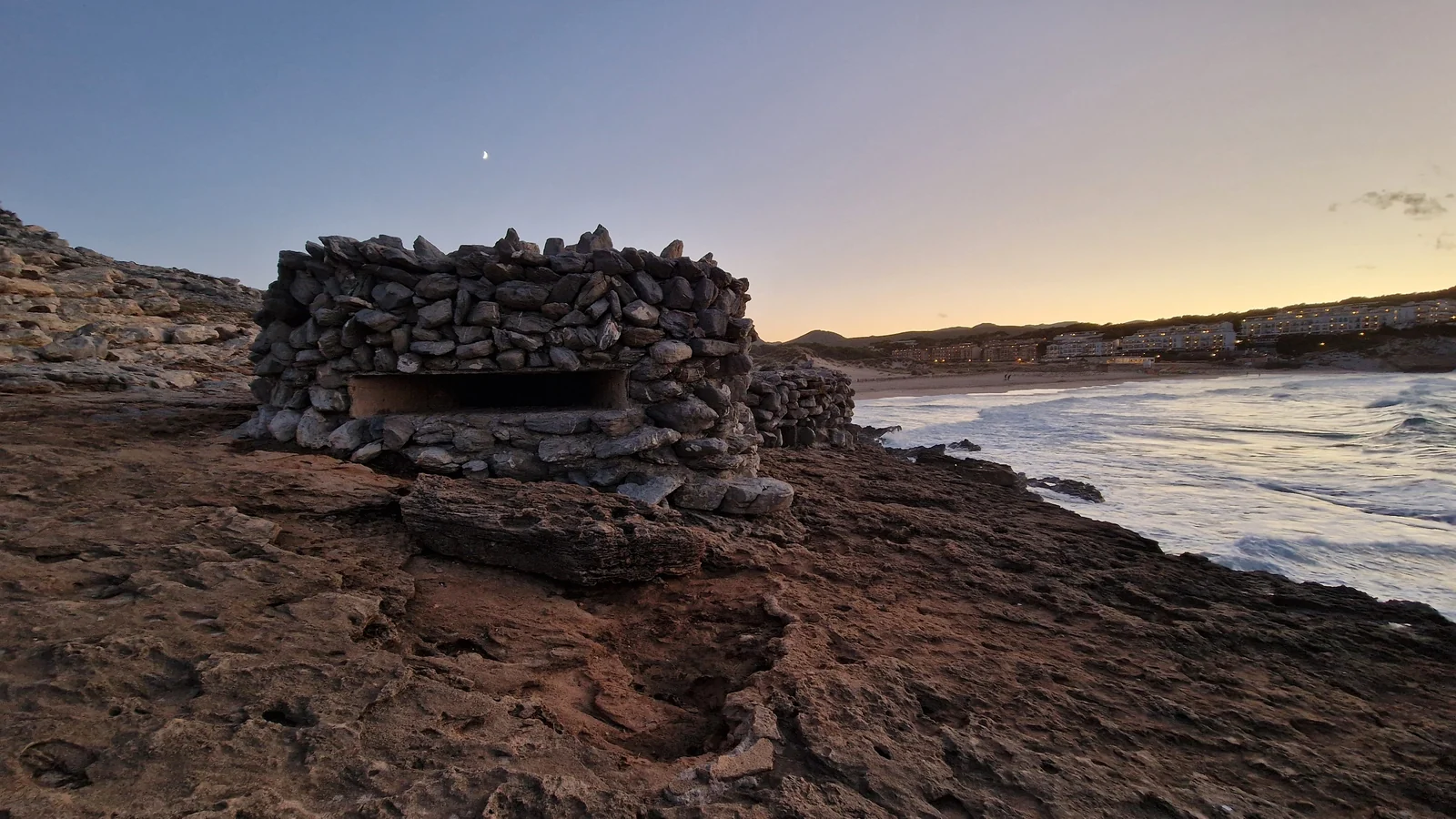 Steinhütte am Fels der Cala Mesquida auf Mallorca, rechts die Bucht mit Sandstrand und Brandung in der Abenddämmerung, September 2024
